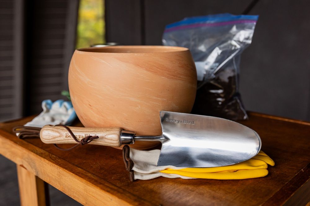 Wooden pot, trowel, yellow gloves, and soil bag on a table for gardening.
