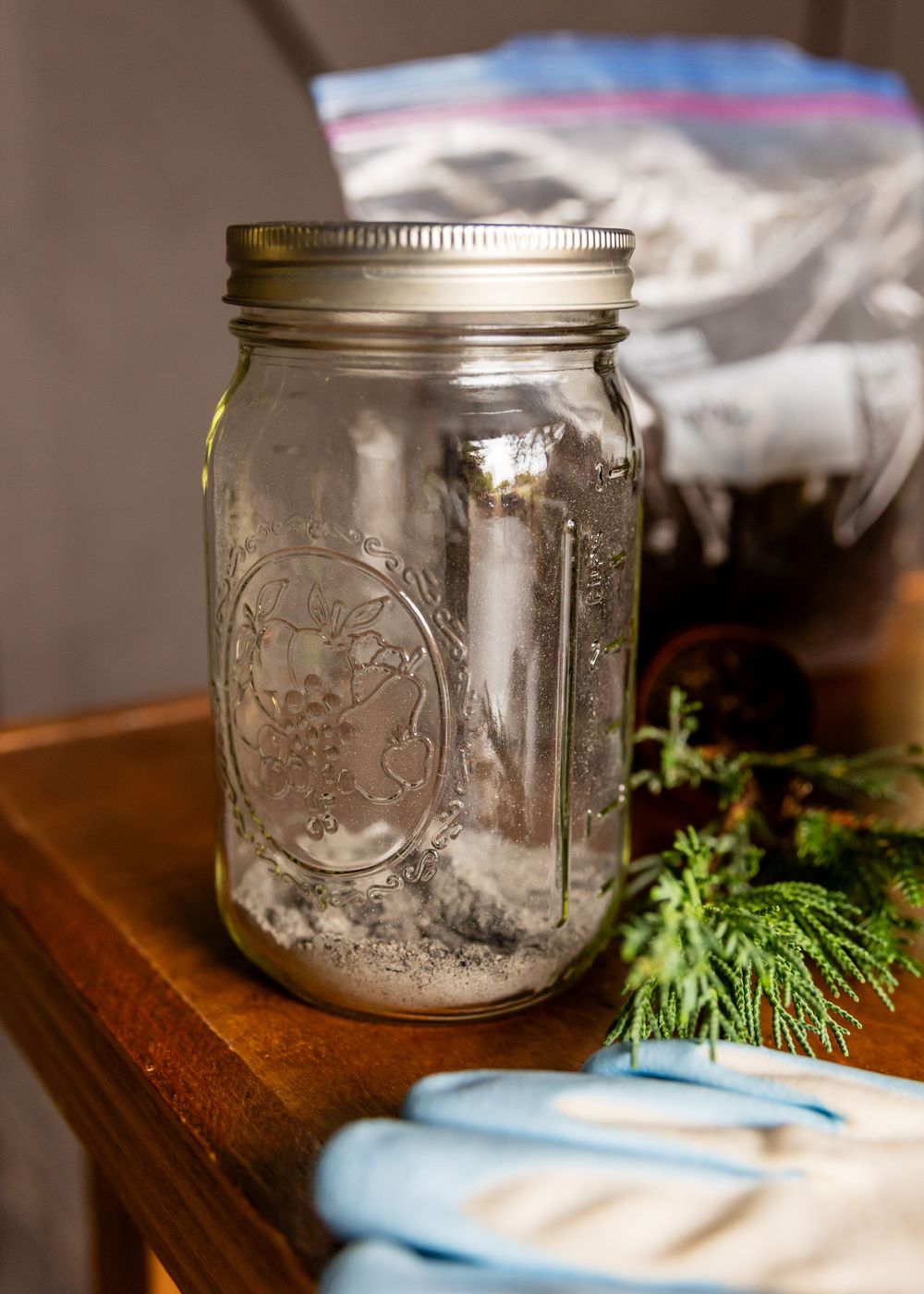 A sealed Ball mason jar with silver lid sits on a wooden table beside evergreen sprigs and blue gloves.