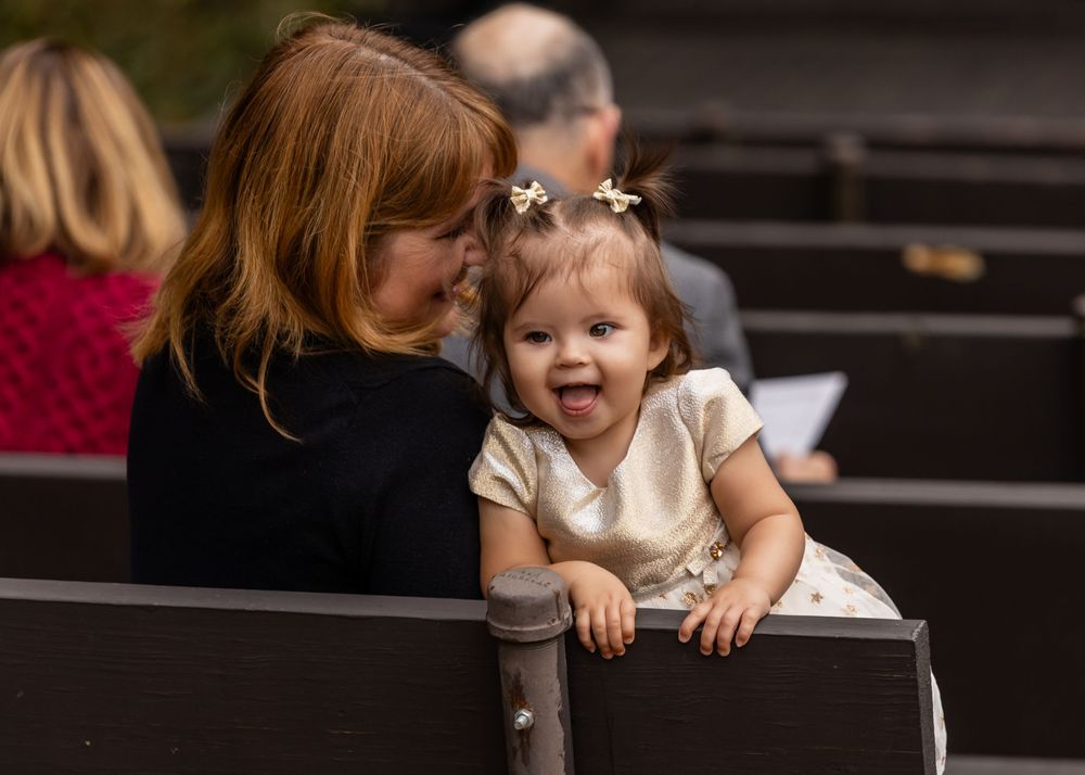 A joyful baby with flower hair clips smiles while sitting on a church pew with a loving red-haired woman at Platte River State Amphitheater