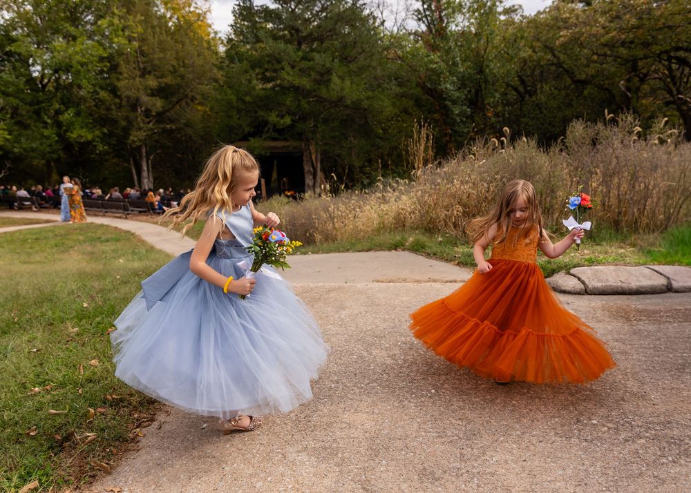 Two flower girls in blue and orange tulle dresses twirl on a garden path during an outdoor wedding ceremony  at Platte River State Park