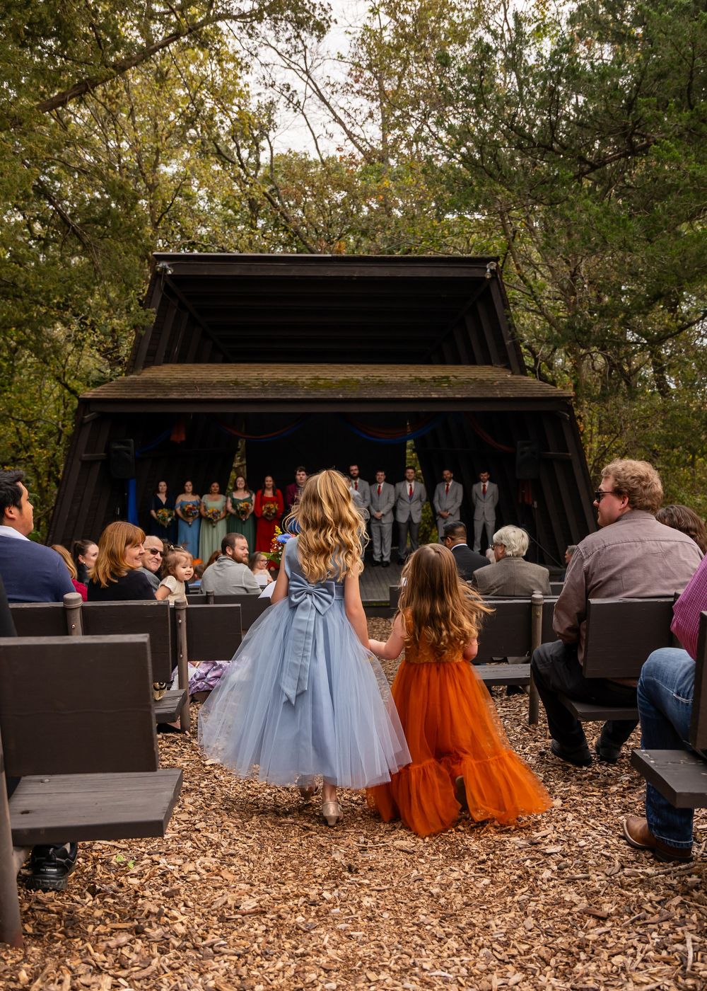 Two flower girls in blue and orange dresses hold hands at an outdoor woodland wedding ceremony stage  at Platte River State Amphitheater