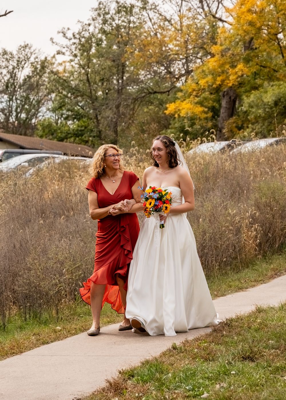 Bride in white gown walks with woman in red dress on autumn pathway holding colorful sunflower bouquet  at Platte River State Park.