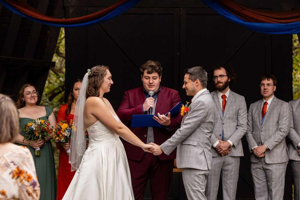 Bride & groom hold hands during outdoor wedding ceremony on wooden stage with wedding party behind them  at Platte River State Amphitheater