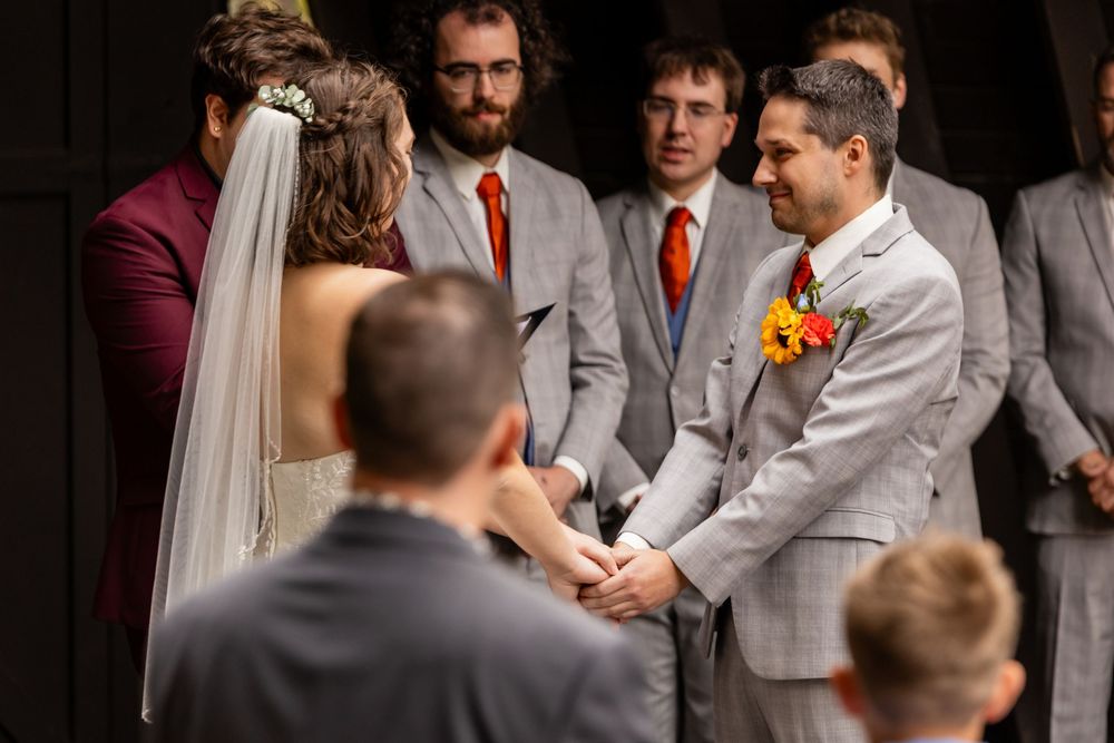 Groom in gray suit with orange tie holds hands with bride at outdoor wedding ceremony  at Platte River State Park Amphitheater