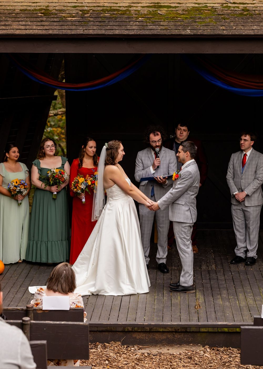 Wedding ceremony on rustic outdoor stage with bridesmaids in green and red dresses and groomsmen in gray suits at Platte River State Park