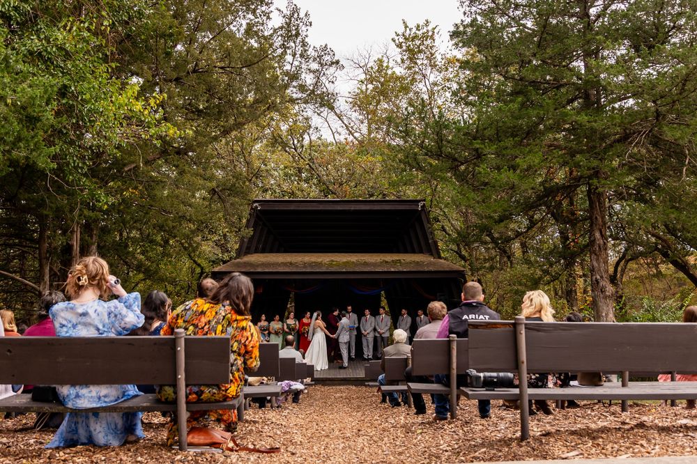 Outdoor wedding ceremony on wooded park stage with guests seated on benches amid autumn trees at Platte River State Park Amphitheater