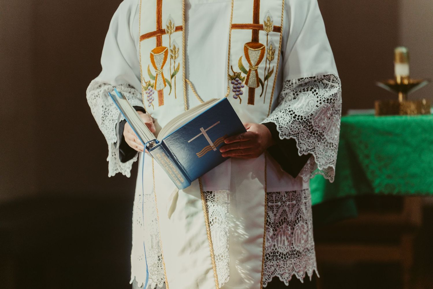 Priest in white vestments with gold embroidery holds a blue Bible during a religious ceremony.