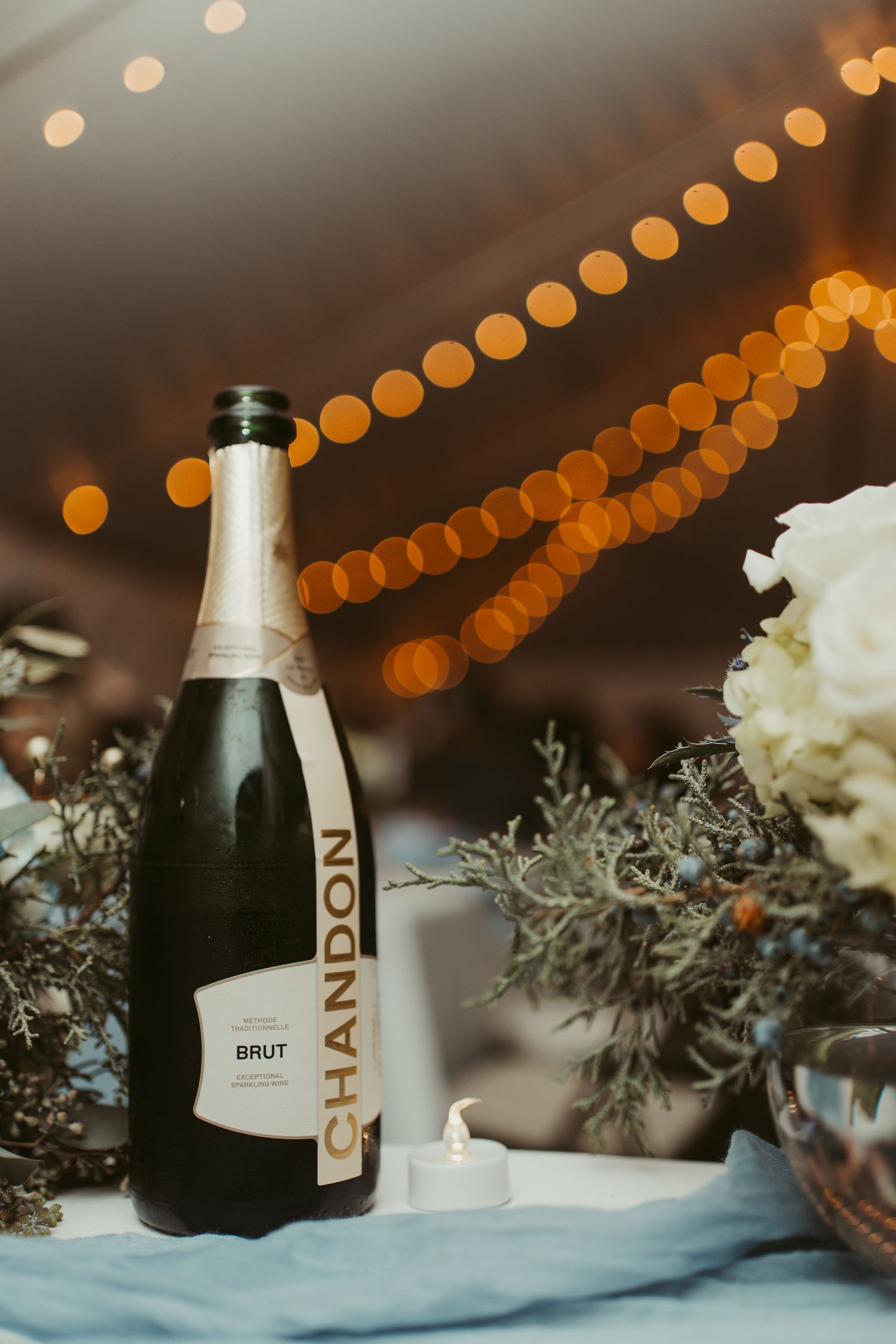 Chandon Brut champagne bottle on a wedding table with white florals, greenery, and bokeh string lights.
