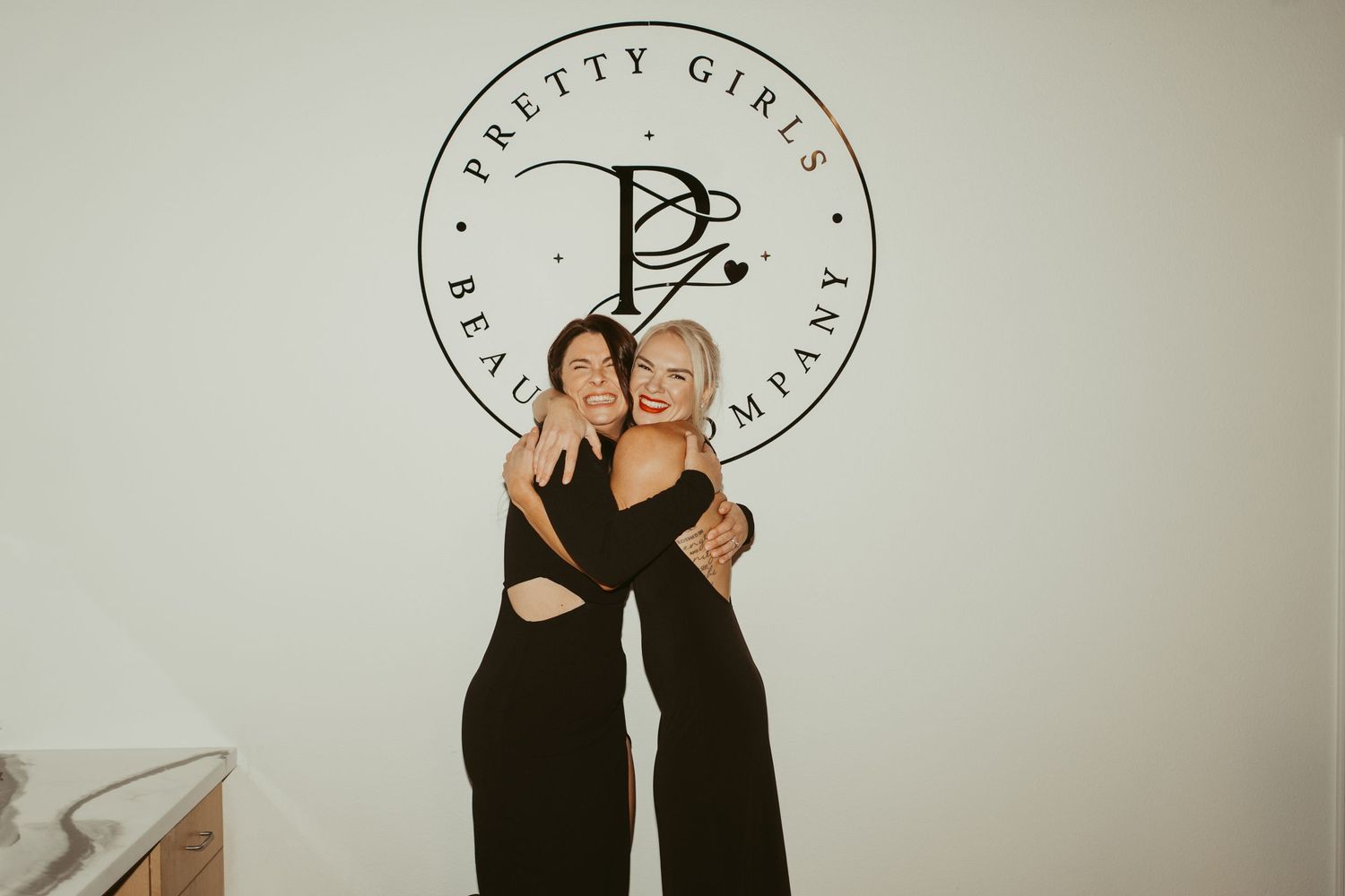 Two women in black outfits smiling and hugging in front of Pretty Girls Beauty Company logo.