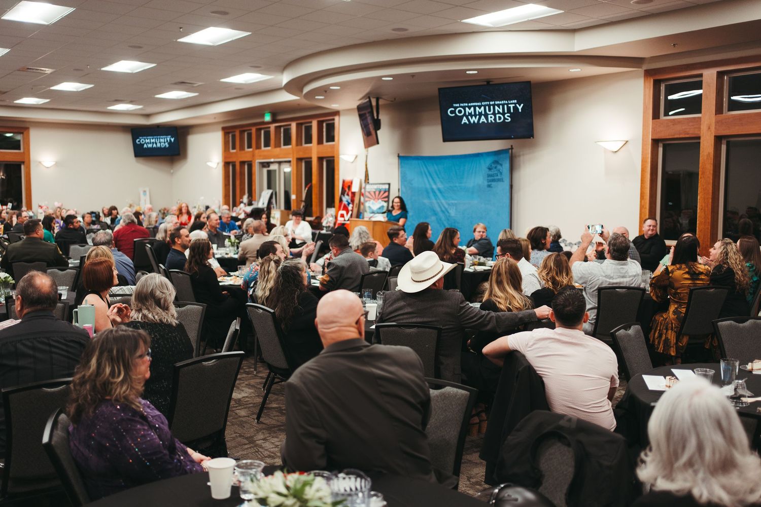 A packed banquet hall hosts a Community Awards ceremony with guests seated at round tables facing a podium.