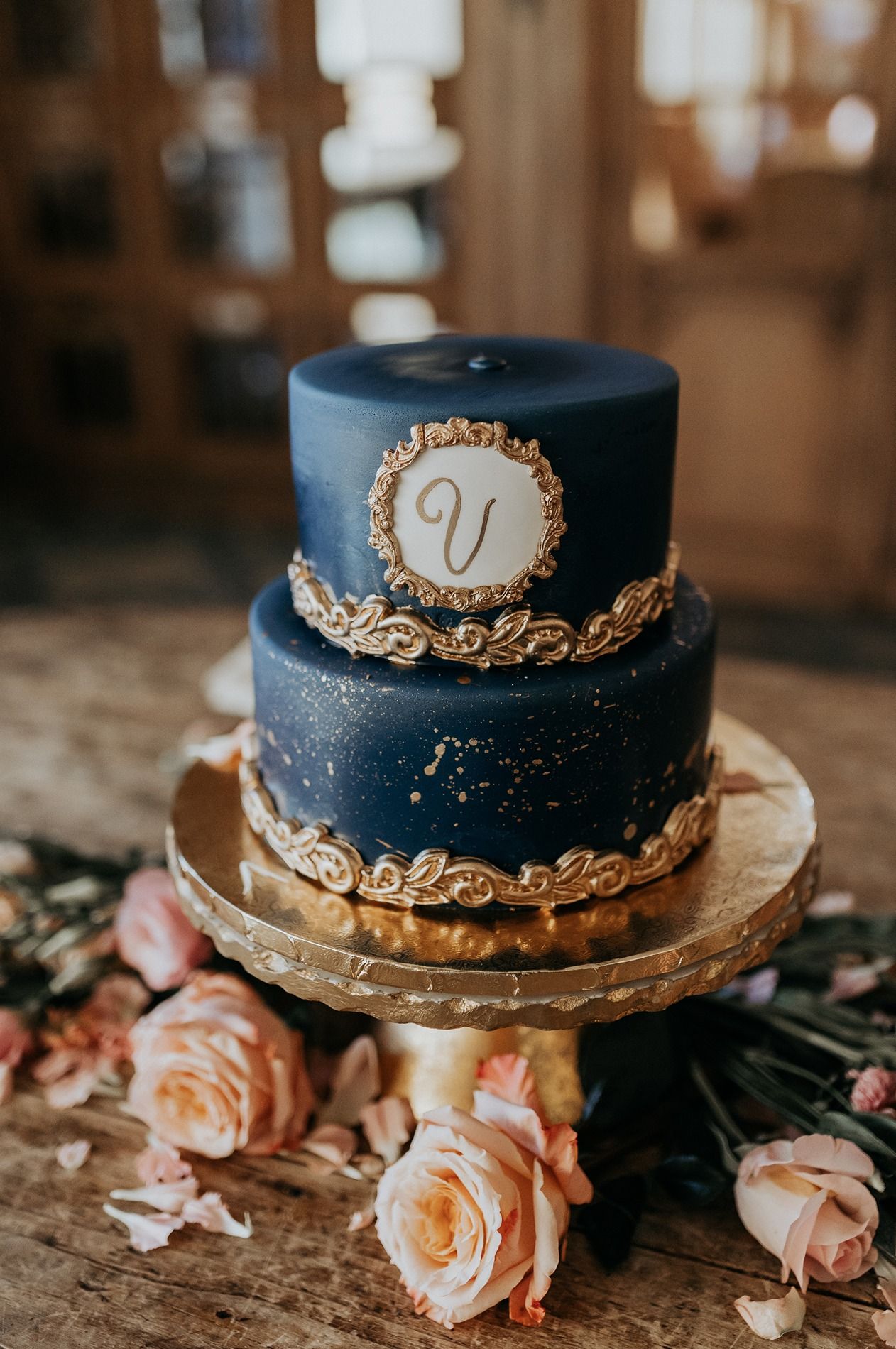 Elegant two-tier navy blue wedding cake with gold trim and monogram V initial, decorated with pink roses on a gold stand.