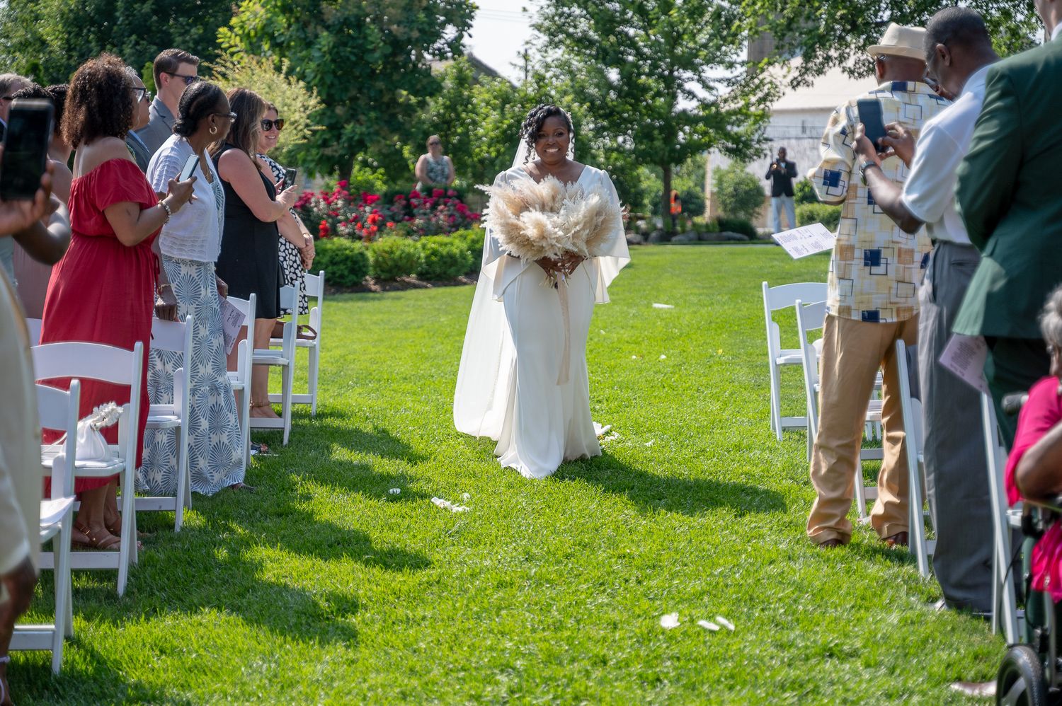 Bride walks down outdoor aisle in white gown holding bouquet, guests seated on white chairs on green lawn.