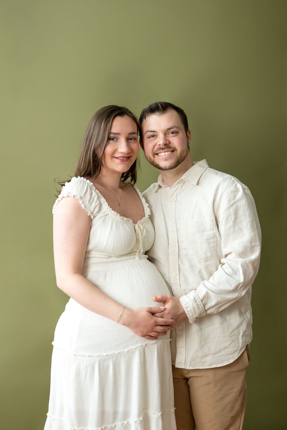 Smiling pregnant couple in white and cream outfits embrace while expecting mother shows baby bump on green background.