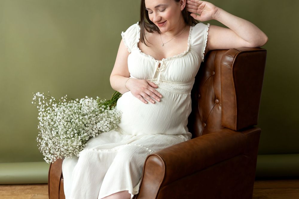 Pregnant woman in white dress seated in brown leather chair, lovingly cradling baby bump with floral bouquet nearby.