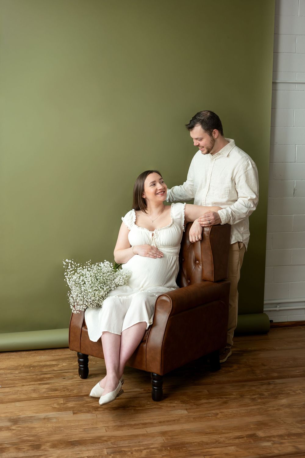 Smiling pregnant couple sharing joyful moment, woman seated in leather chair as man stands beside her smiling.