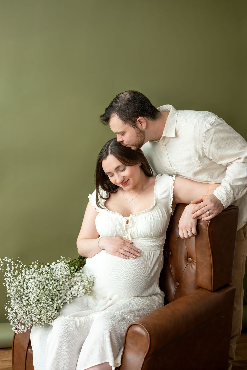 Man tenderly kissing pregnant woman's head as she sits in leather chair cradling bump, white flowers nearby.