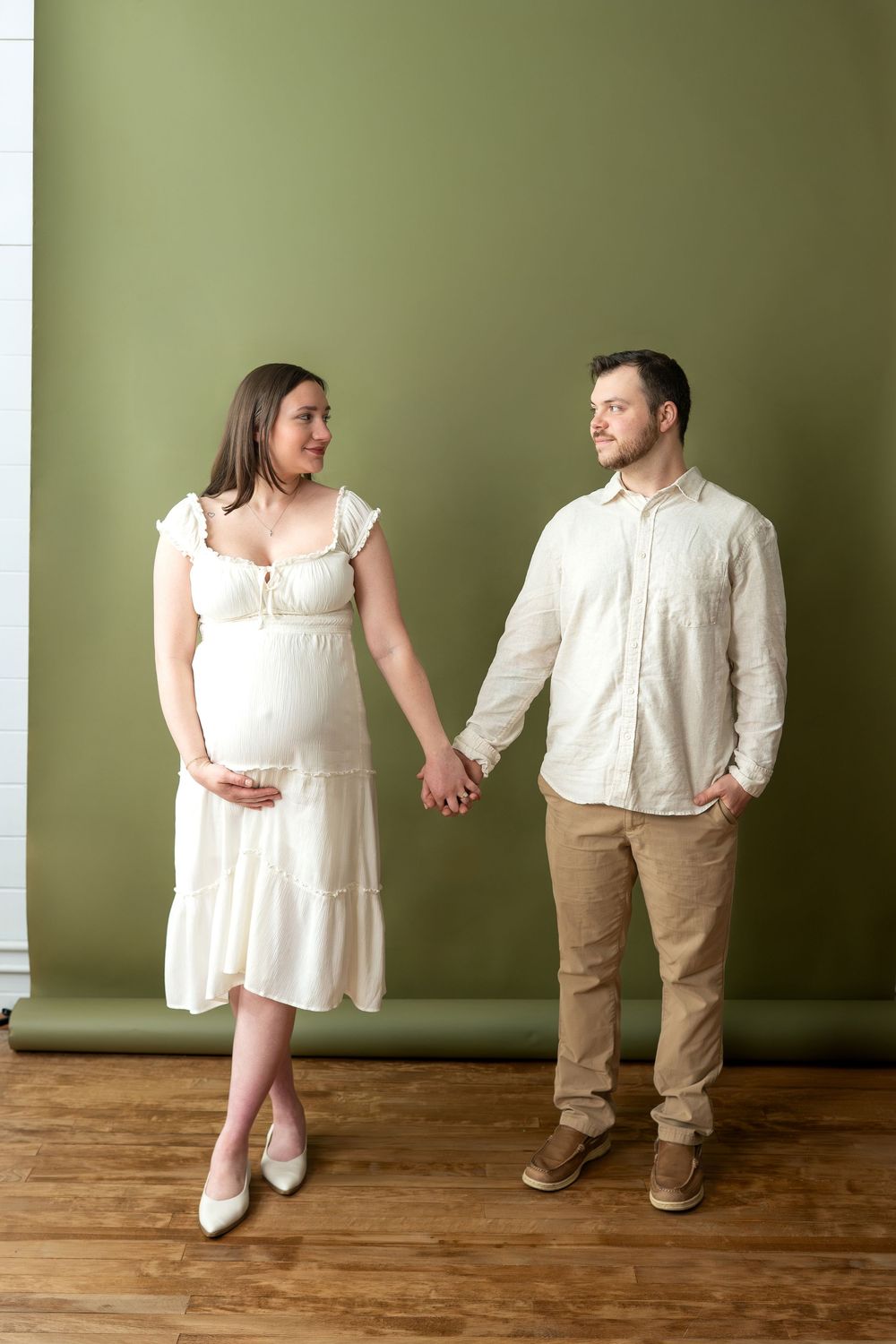 Pregnant couple holding hands and smiling at each other, woman in white dress against olive green wall.