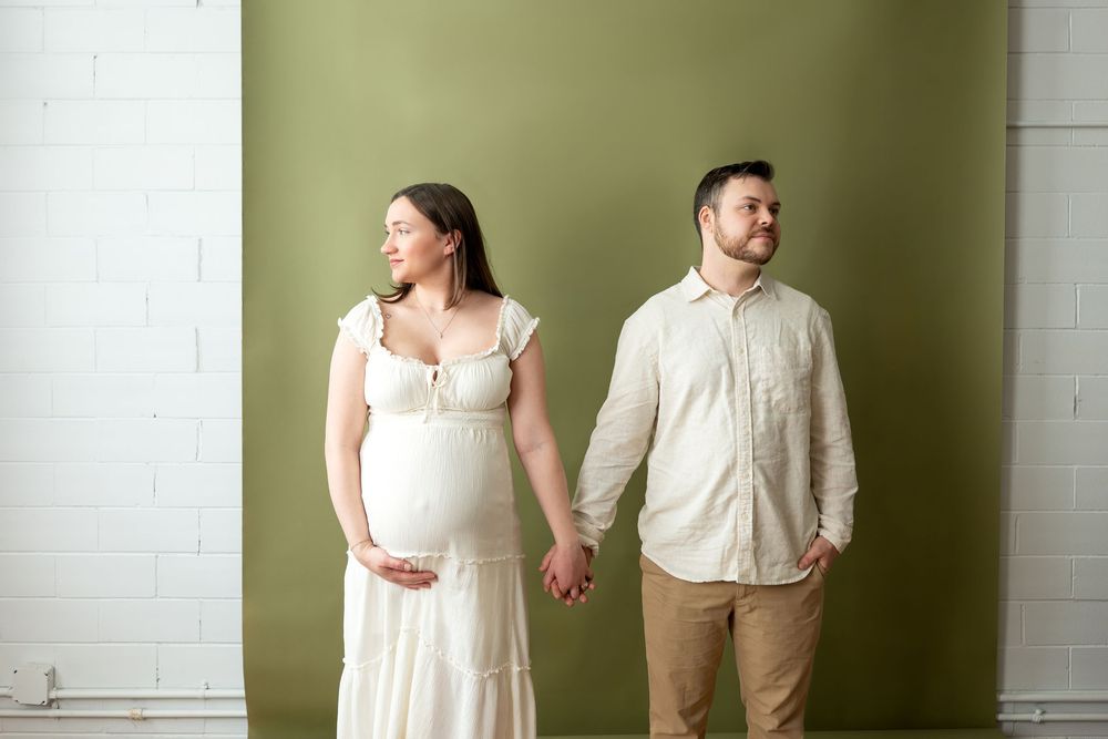 Pregnant couple holding hands while standing against olive green wall, woman in white dress cradling baby bump.