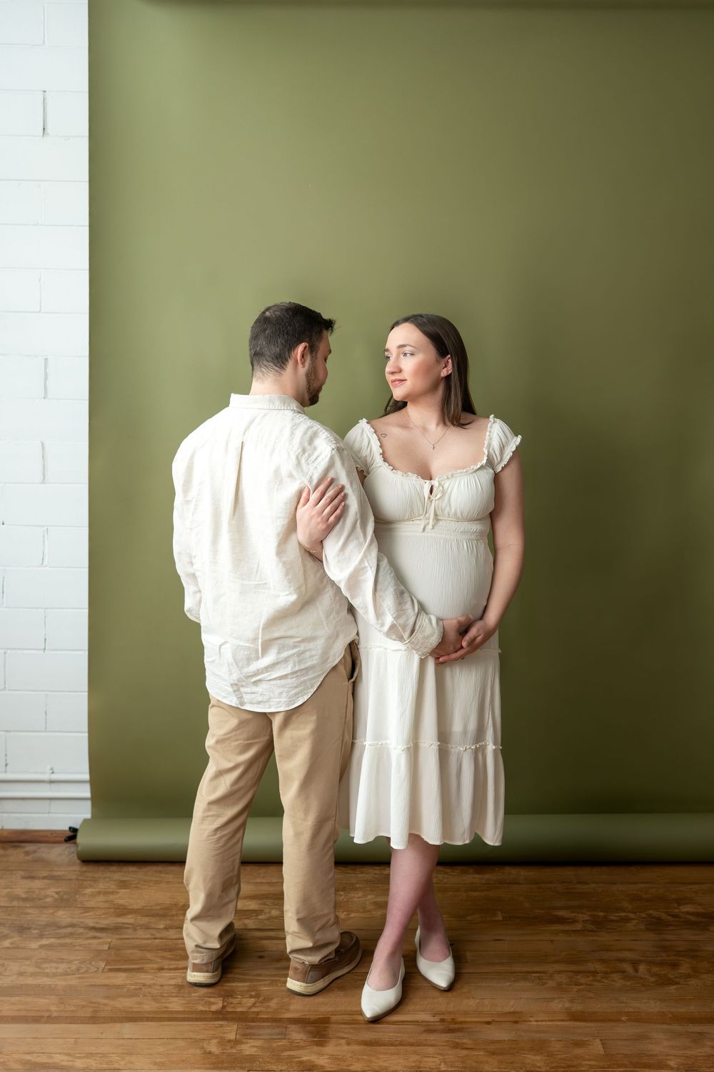 Expectant couple in white outfits share a tender moment against an olive green studio wall.