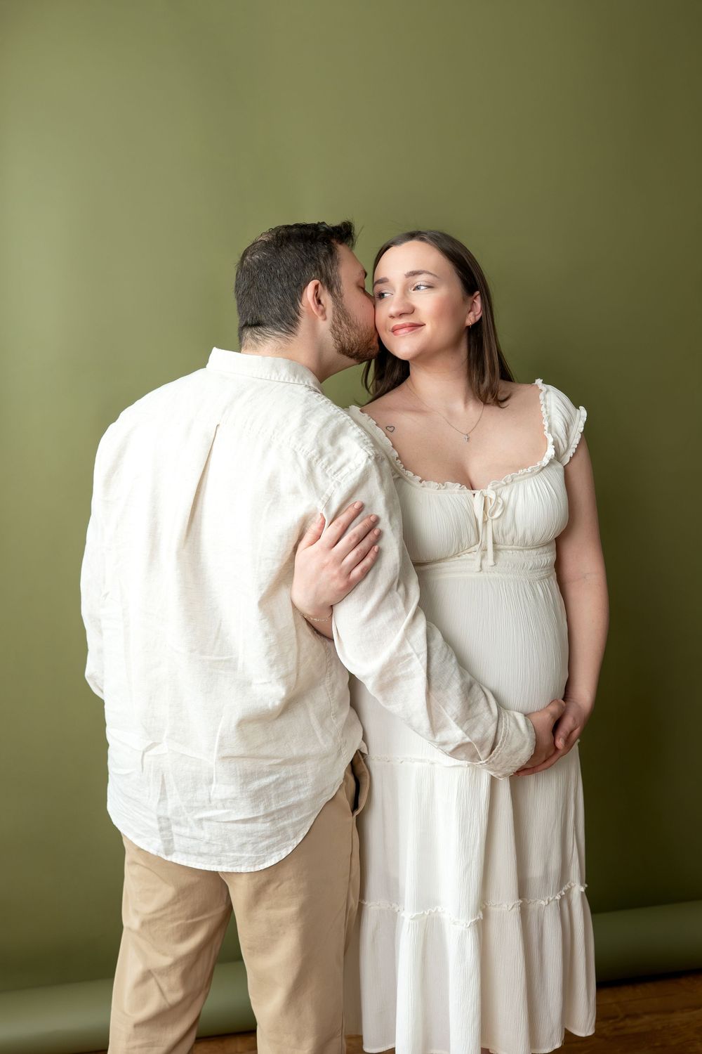 Man kisses pregnant woman's cheek as she smiles, both dressed in white against green backdrop.