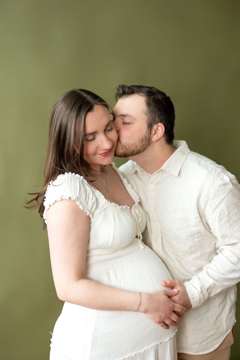 Man tenderly kisses pregnant partner on cheek while holding her baby bump in white maternity studio portrait.