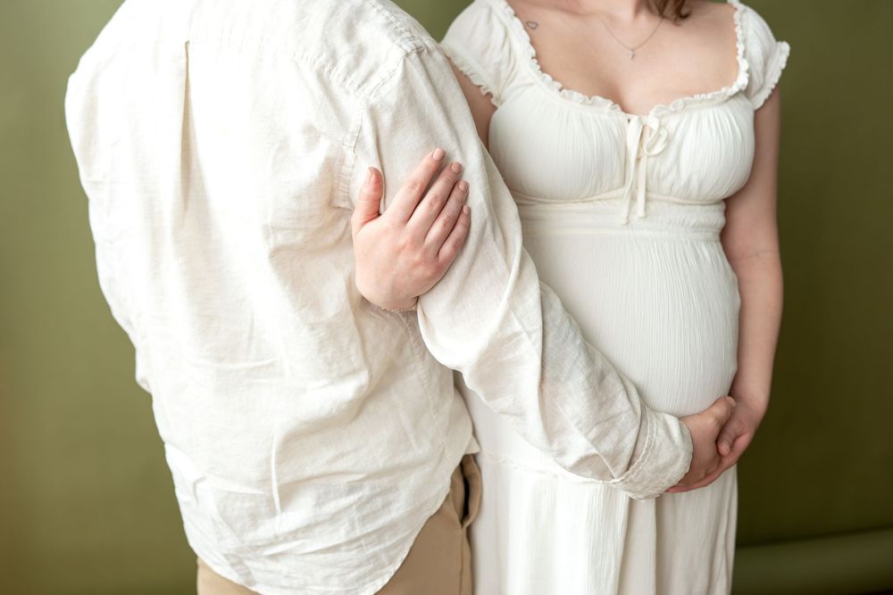 Close-up of pregnant woman in white dress with partner's hand on her back, green background.