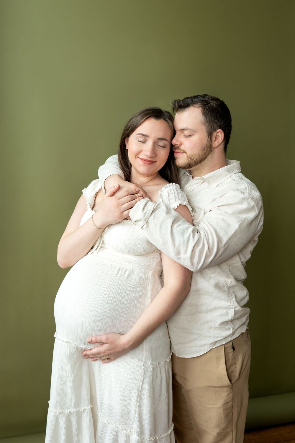 Couple embraces with eyes closed, pregnant woman in white dress cradled by partner, green wall.