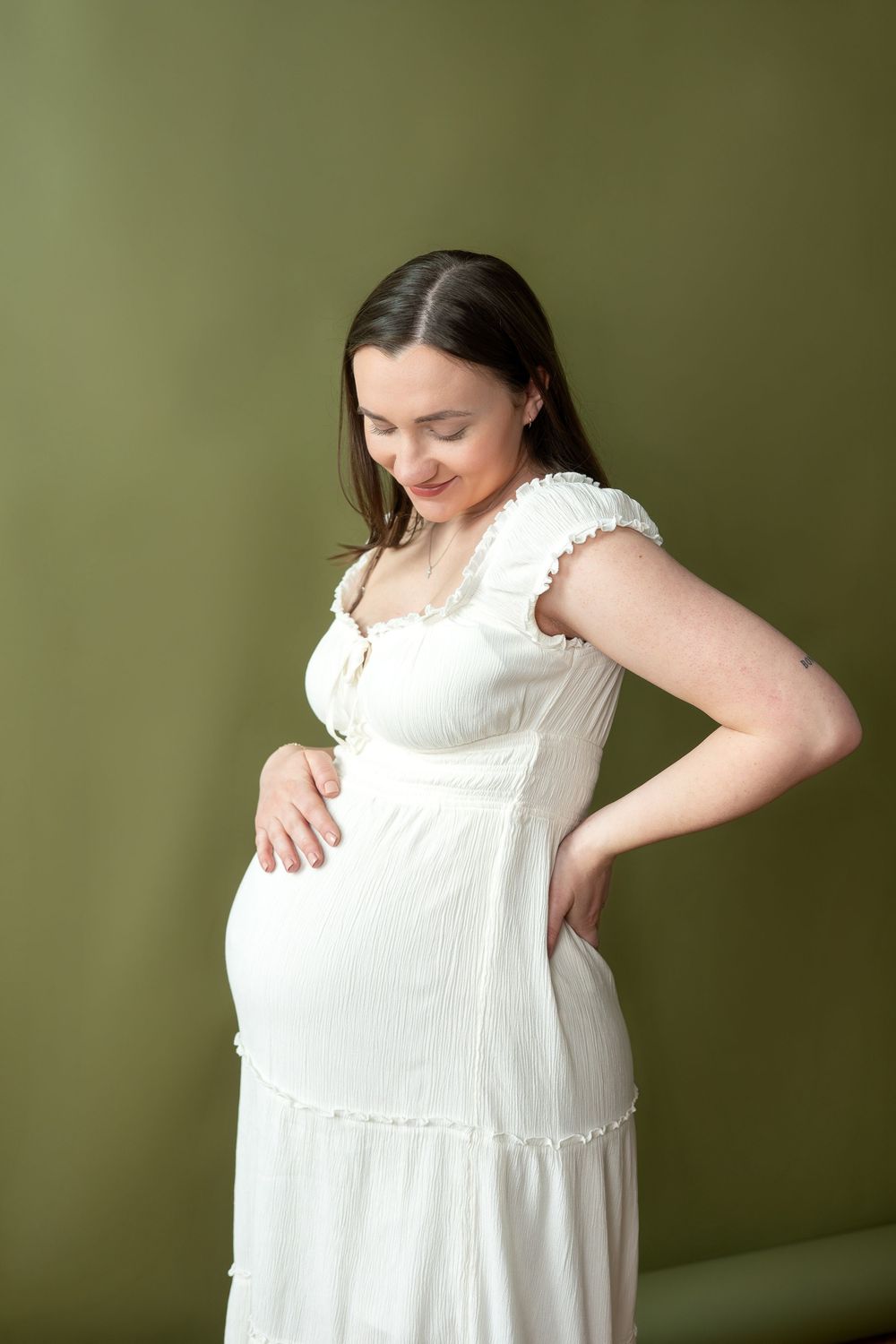 Pregnant woman in white dress looks down at baby bump, smiling softly against green backdrop.