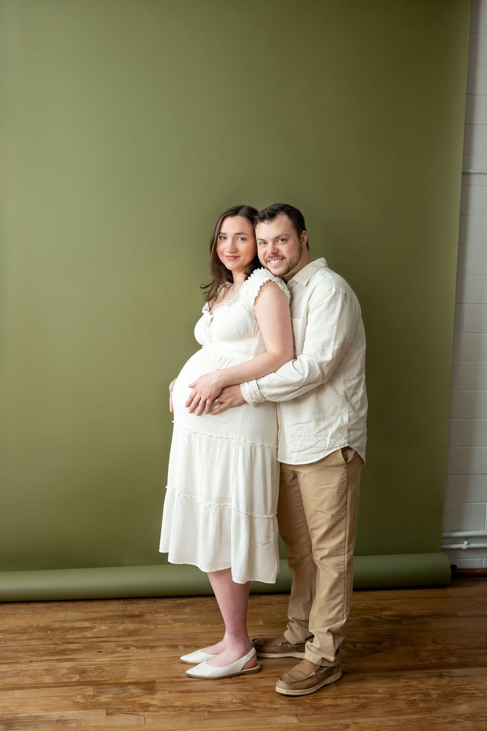Expectant couple poses together with man hugging pregnant woman from behind against olive green studio wall.