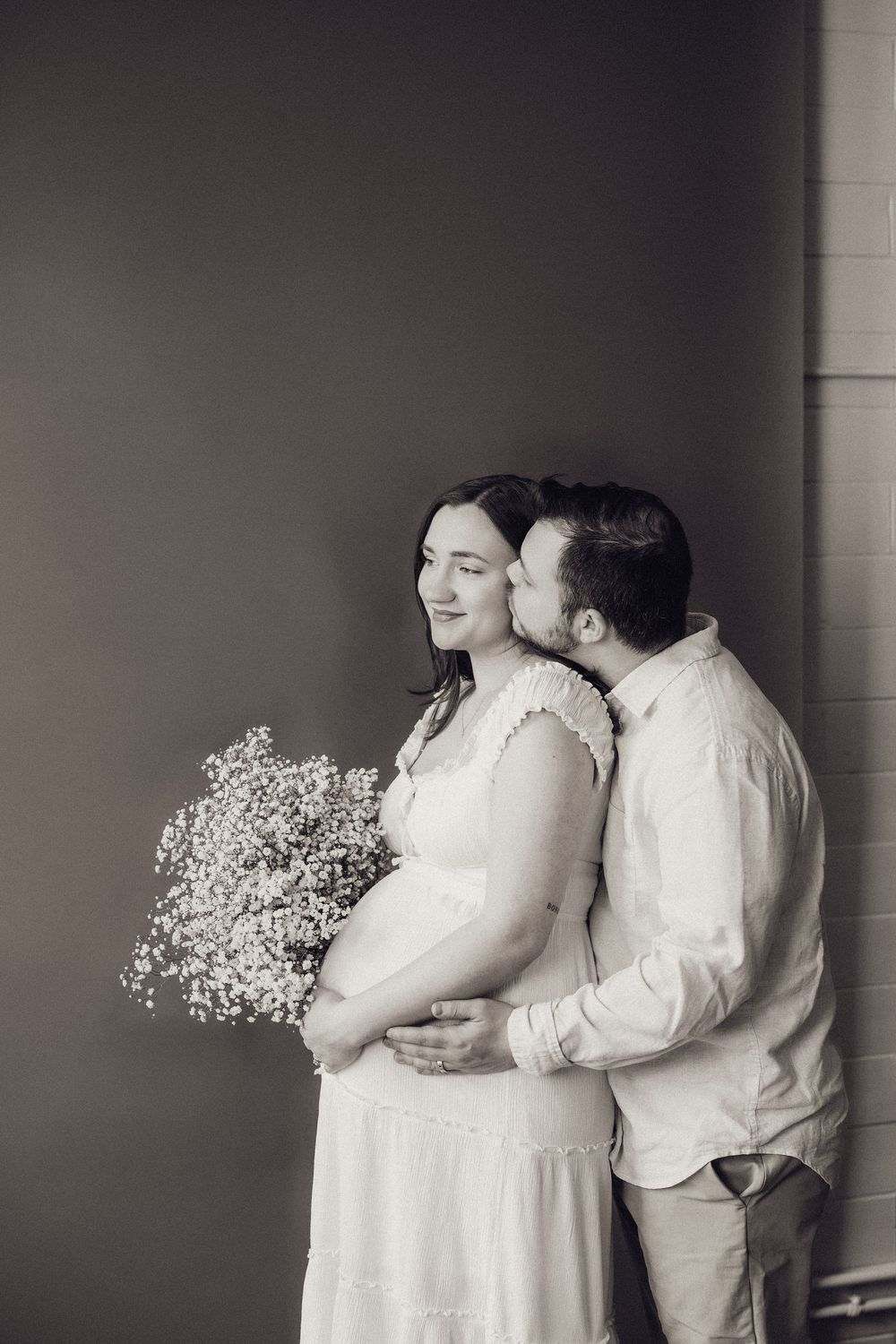 Black and white photo of couple, man kissing pregnant woman holding flowers in white dress.