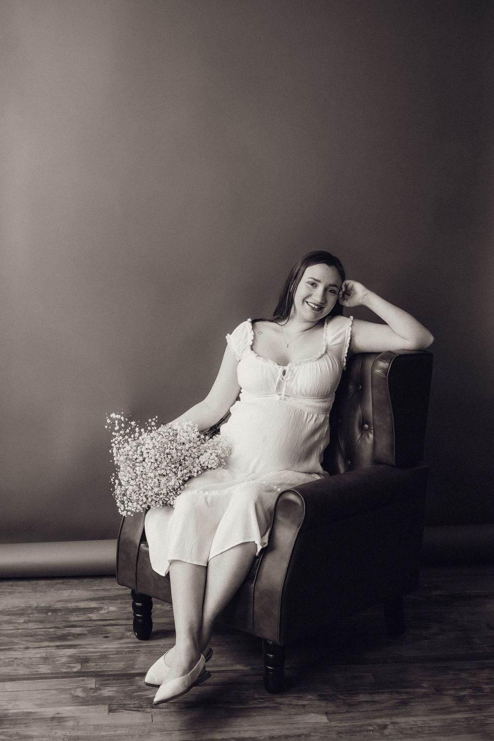Black and white photo of smiling pregnant woman in white dress sitting in leather armchair with flowers.