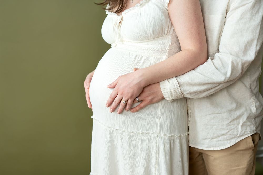 Close-up of pregnant woman in white dress with partner embracing her baby bump against olive green background.