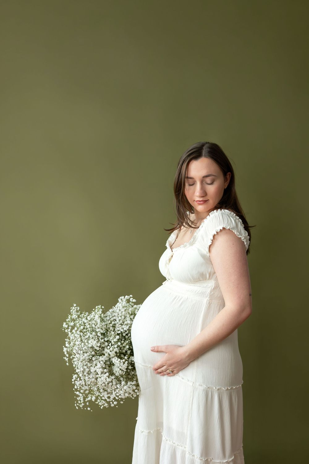 Pregnant woman in white dress cradles baby bump while holding white baby's breath flowers on green background.