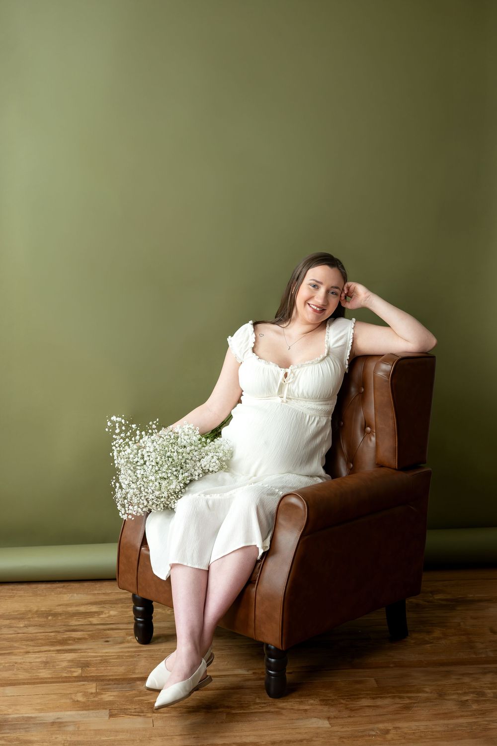 Smiling pregnant woman in white dress sitting relaxed in brown leather chair with baby's breath bouquet.