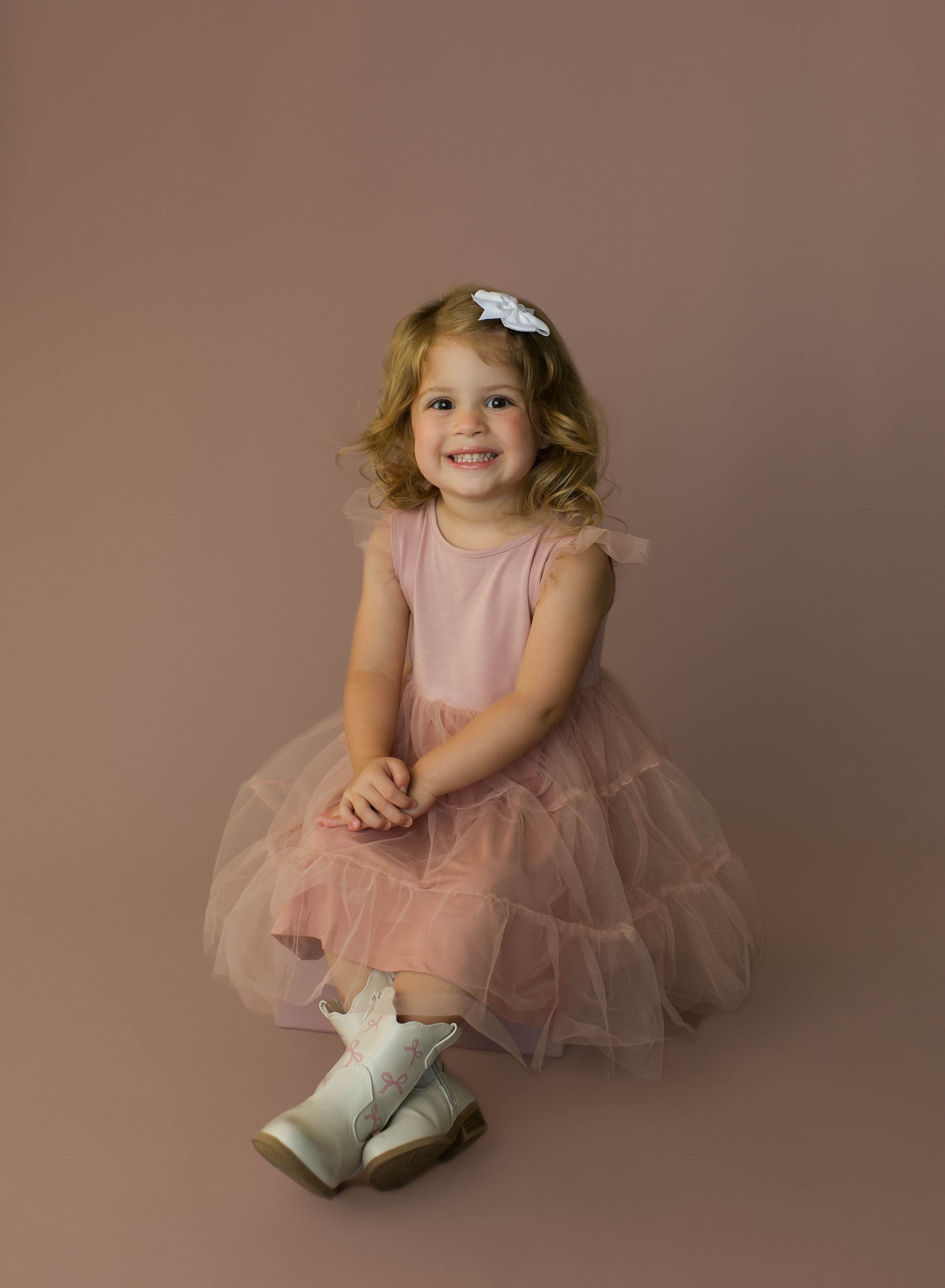 A smiling toddler girl in a pink tulle dress and white shoes sits posed against a mauve studio backdrop.