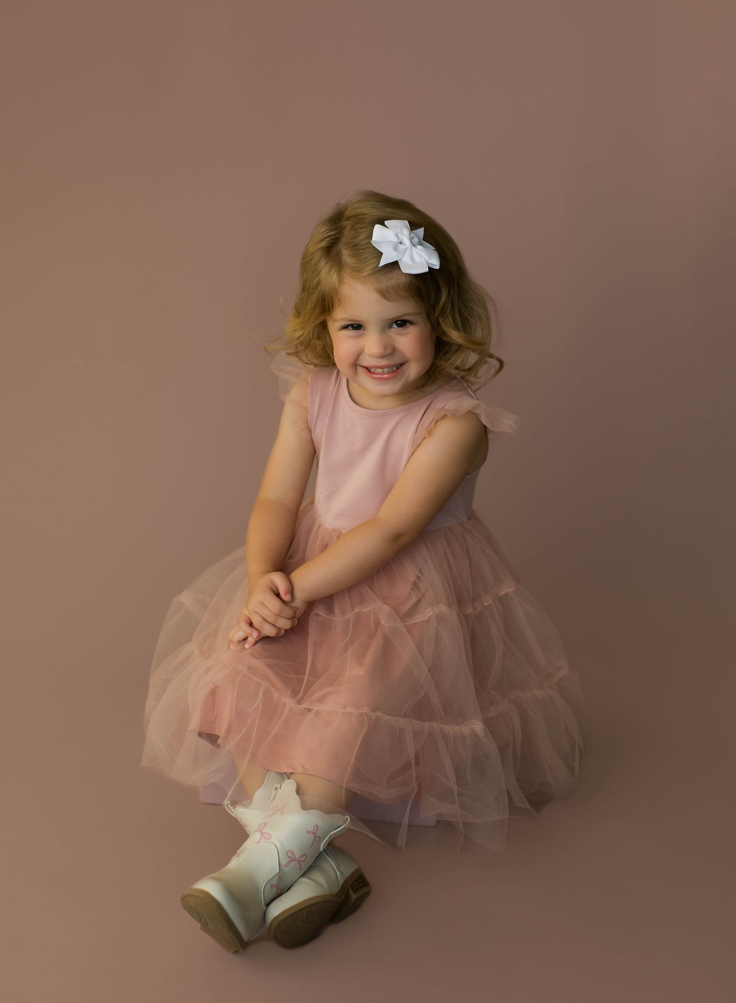 A smiling toddler girl in a pink tulle dress and white bow hairclip poses cheerfully against a mauve studio background.