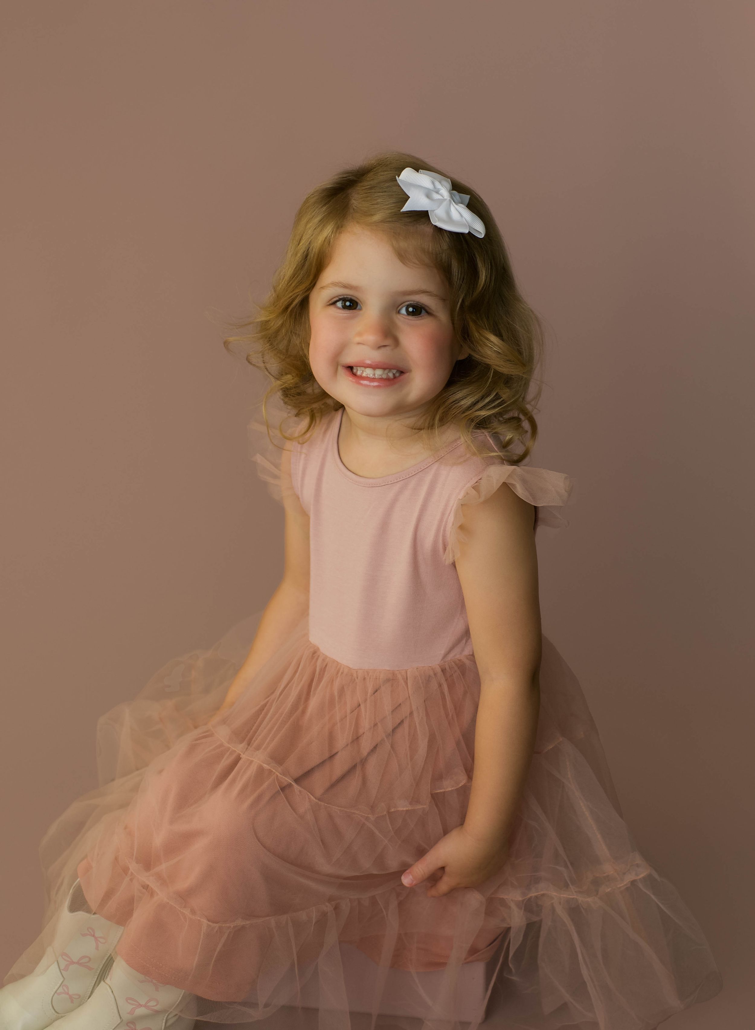 A smiling toddler girl in a pink tulle dress with a white bow hairclip poses for a portrait photo.