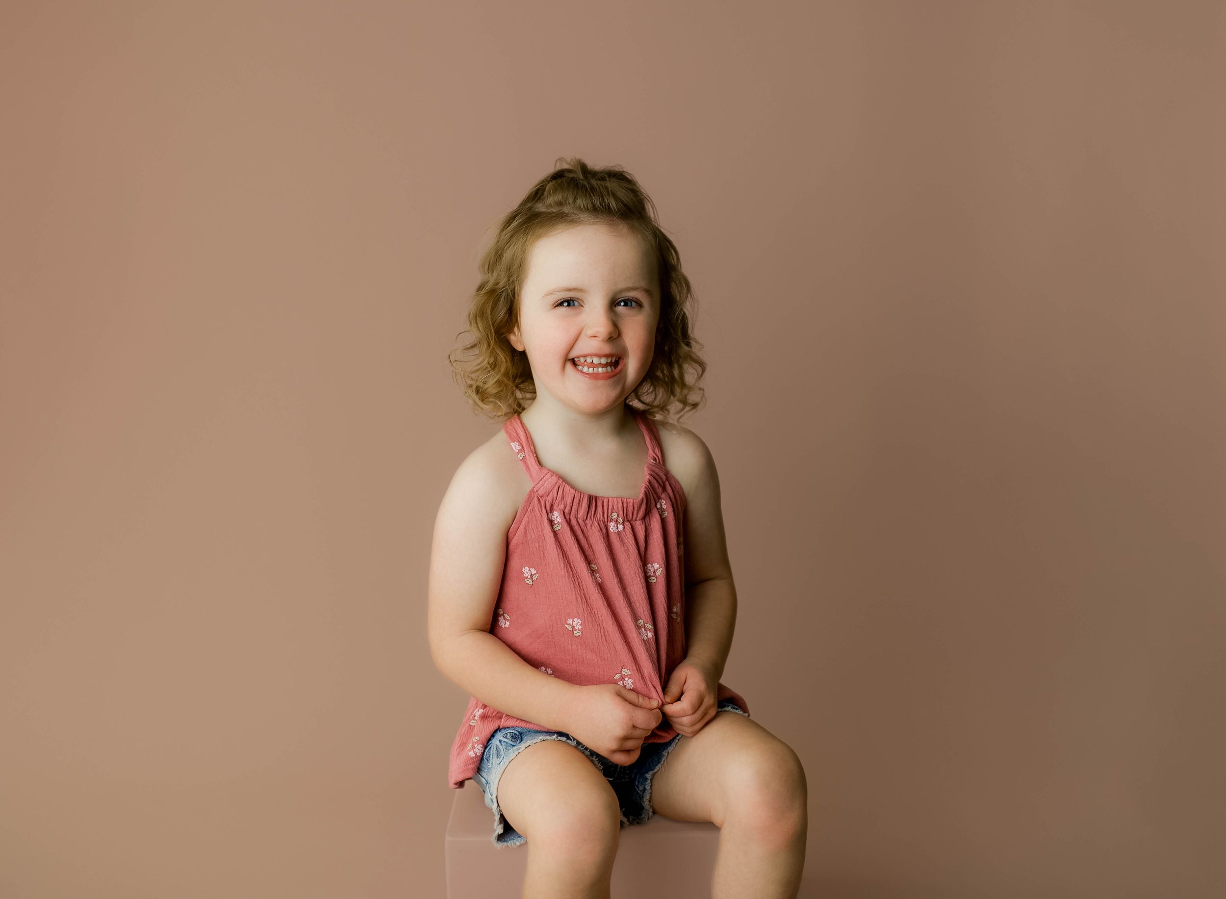 Smiling young girl with curly hair wearing a pink top and denim shorts, sitting against a beige studio background.