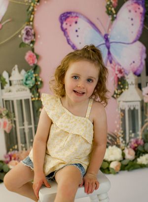 Young girl in yellow floral top and denim shorts smiling in front of a butterfly backdrop with flowers.