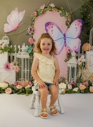 Young girl smiling in yellow floral top seated before a colorful butterfly garden backdrop.
