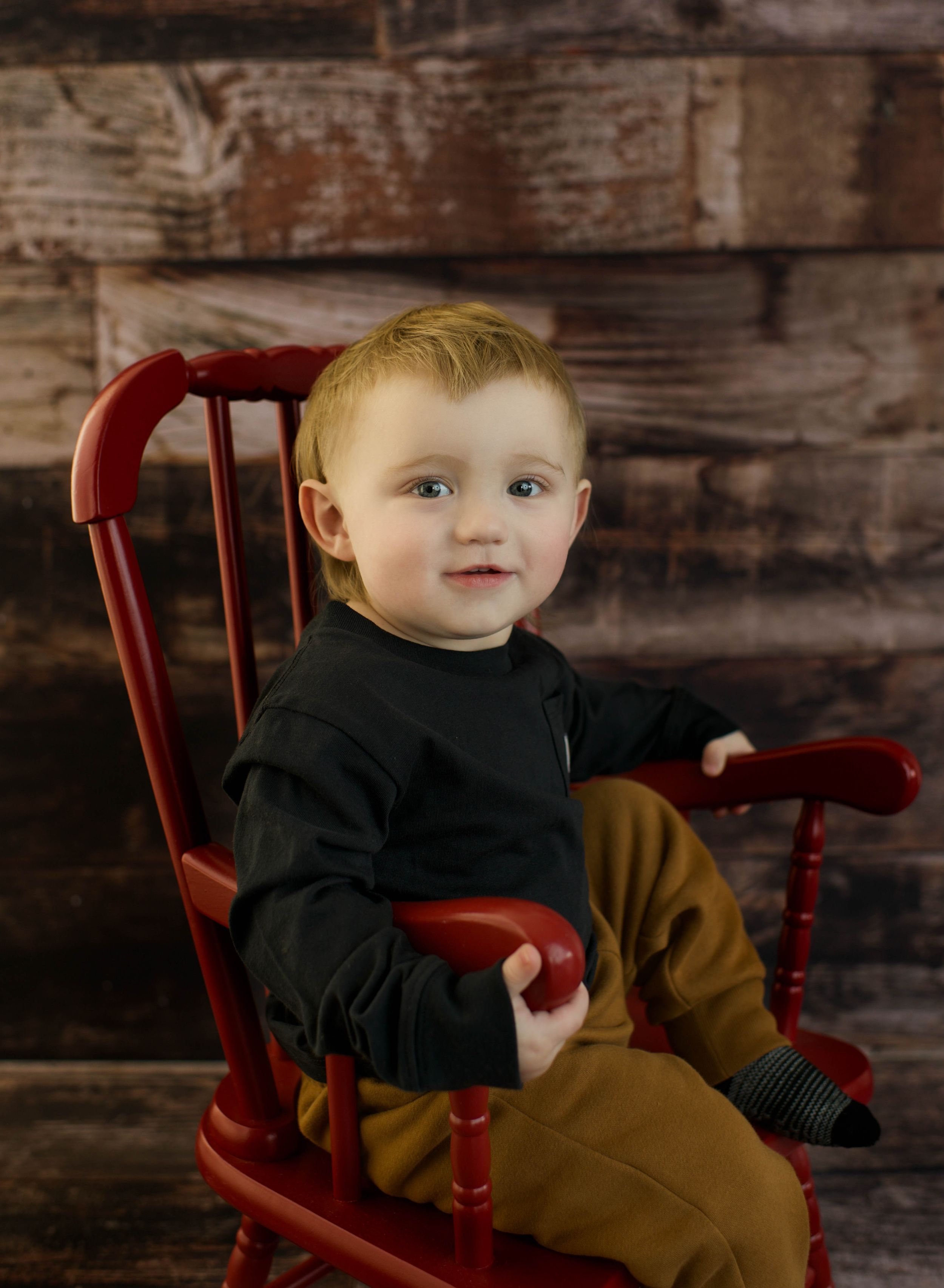 Toddler boy with blonde hair sitting in a red wooden rocking chair against a rustic wood backdrop.