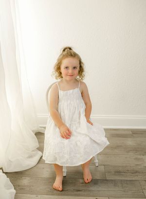 Young girl in white sundress standing barefoot on wood floor near sheer curtains in bright airy room.