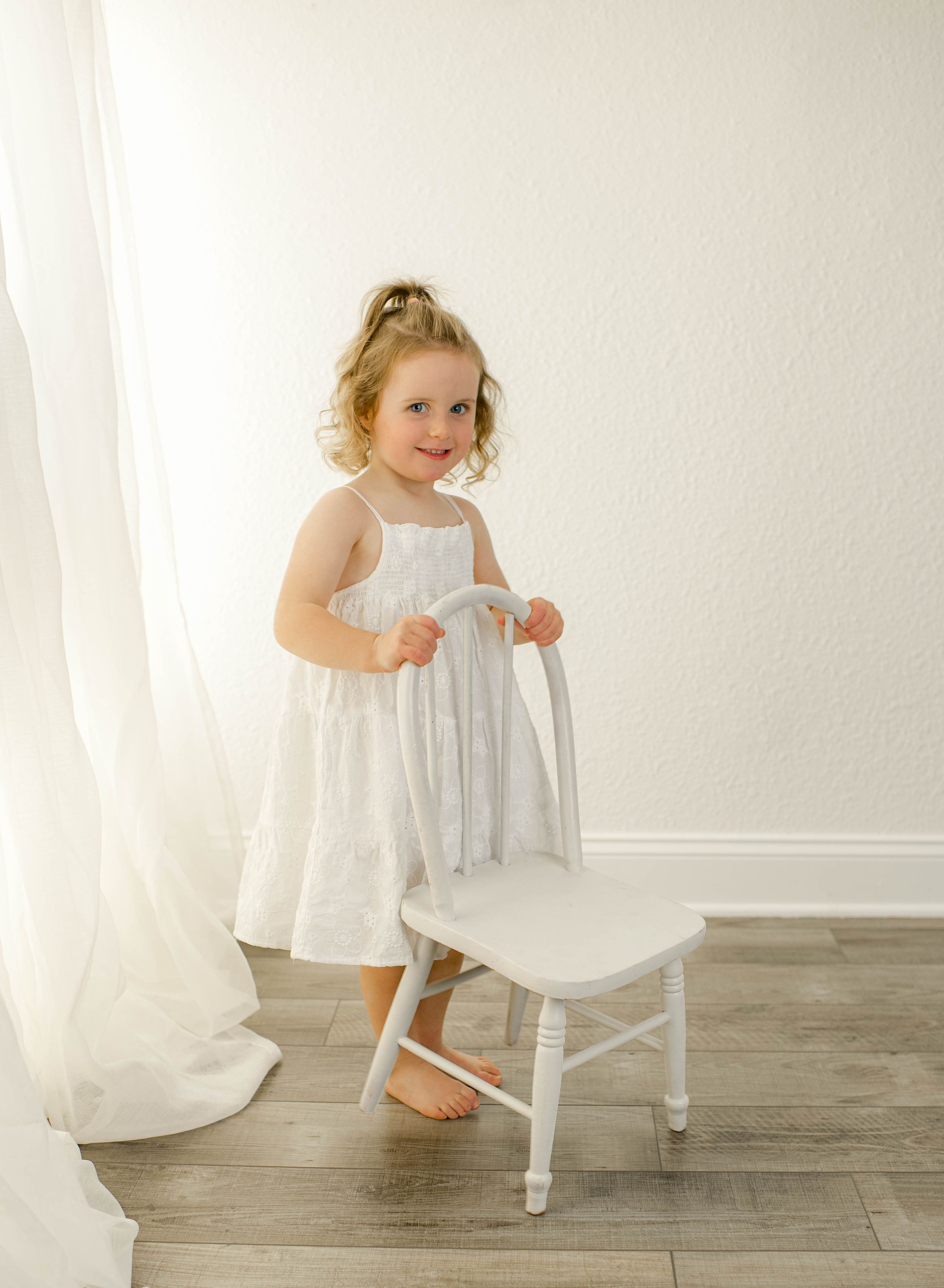 Young girl in white dress standing barefoot beside a small white chair in a bright, airy room.