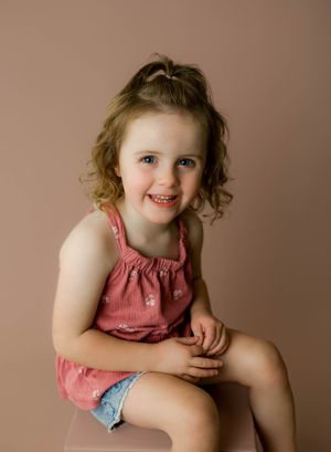 A smiling young girl with curly hair sits wearing a pink top and denim shorts against a warm beige background.