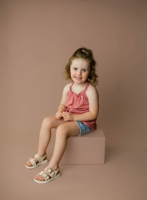 Young girl smiling in pink top and denim shorts sitting on a pink block in a studio portrait.