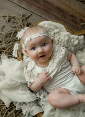 Adorable baby girl in white lace outfit and floral headband nestled in a rustic wooden bowl with daisy accents.