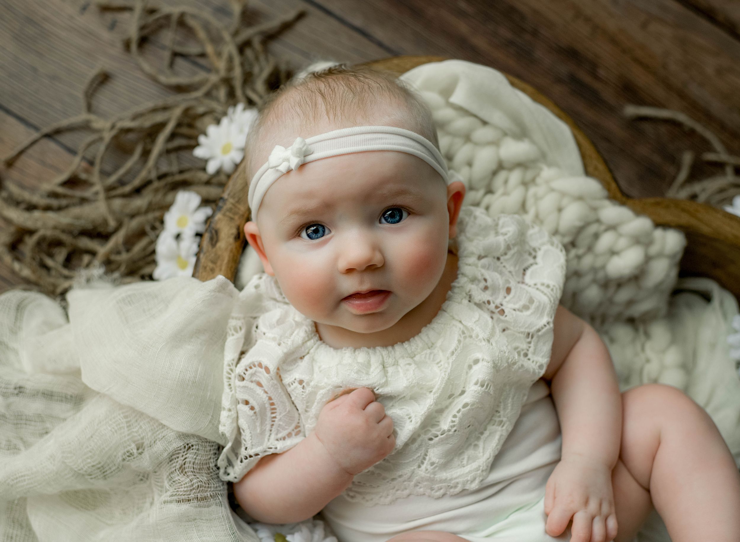 Adorable baby girl in white lace dress and floral headband posed in rustic wooden bowl with soft cream blanket.