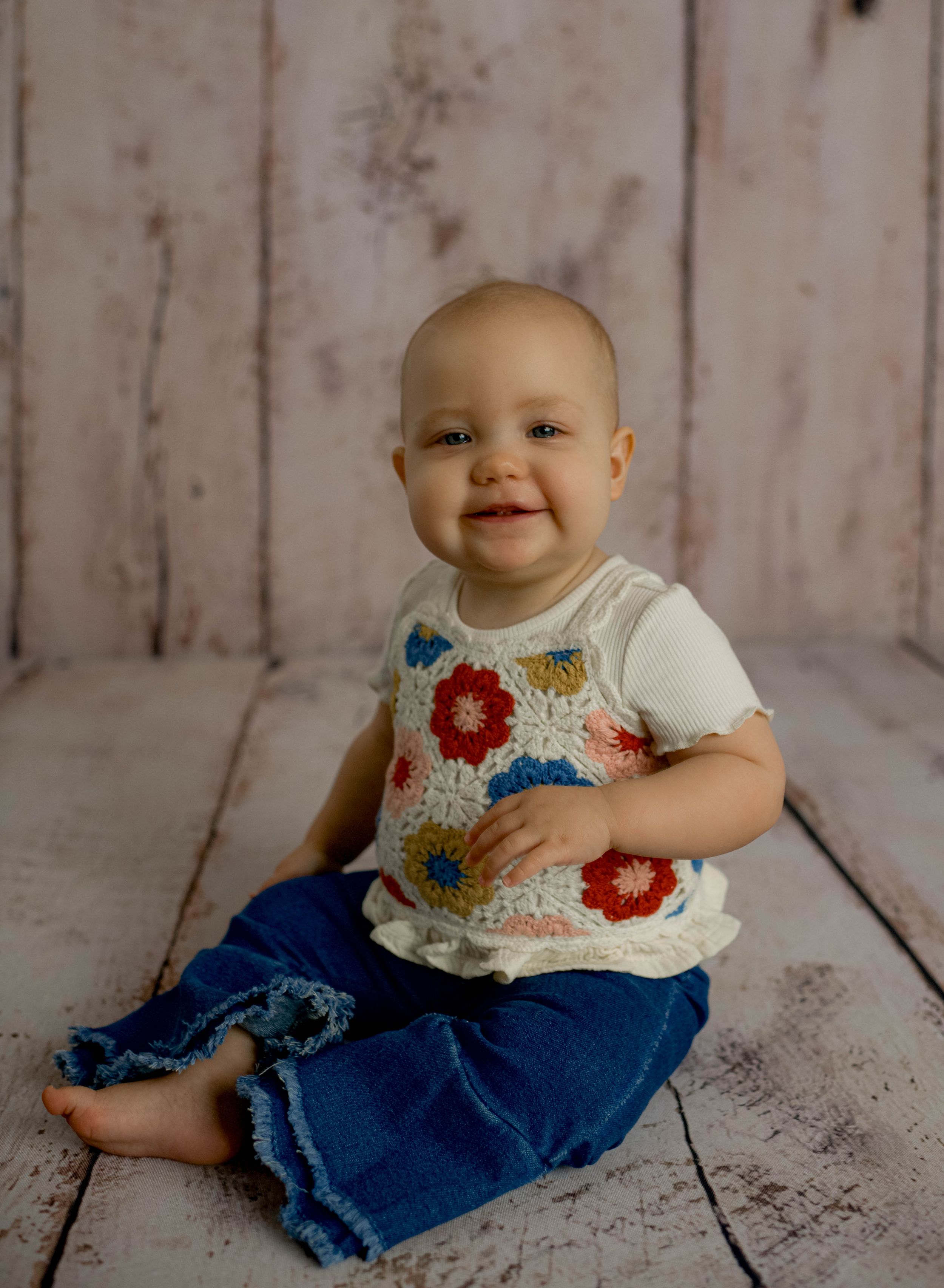 Smiling baby in crochet floral vest and denim pants sitting on white wood floor for a portrait photo.