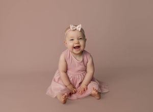 Smiling baby girl in pink dress and bow headband sitting on beige studio backdrop for professional portrait.