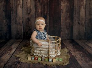 Baby girl in floral outfit sits in rustic basket with alphabet blocks spelling a name on wood floor backdrop.